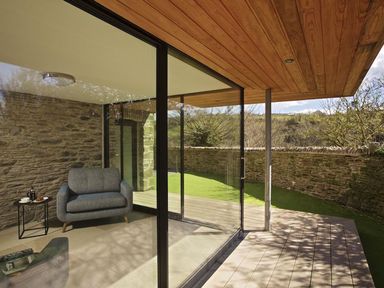 A seating area with a gray armchair and a small round table inside a glass-walled room with a stone wall and a wooden ceiling at The Milking Shed in Bantham