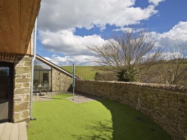 An outdoor area with artificial grass a stone wall patio with table and chairs and a tree at The Milking Shed in Bantham