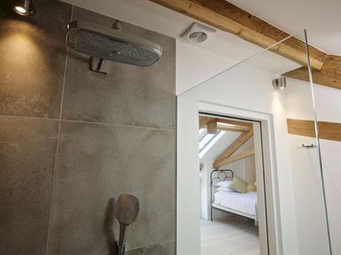 A bathroom shower area with grey tiles and a metal shower head opening into a bedroom with a bed under wooden beams at The Milking Shed in Bantham