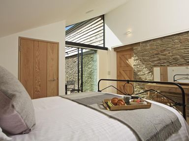 A bedroom with a bed with gray pillows and blanket a wooden door stone wall and large glass window at The Milking Shed in Bantham