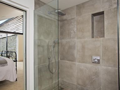 A shower area with beige tiles glass partition and built in niche next to a bedroom with a metal bed frame at The Milking Shed in Bantham