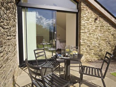 An outdoor patio with a black table and four black chairs in front of stone walls and glass sliding doors at The Milking Shed in Bantham