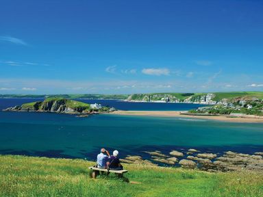 Two people sitting on a bench on a grassy hill overlooking a rocky coastline with clear blue water and a small island with buildings at The Milking Shed in Bantham
