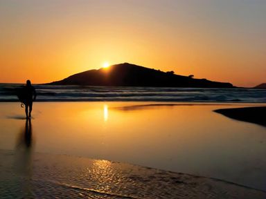 A surfer walking on a beach at sunset with an island in the background at The Milking Shed in Bantham
