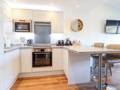 A kitchen with white cabinets a built-in oven a microwave a kettle toasters a coffee machine and a breakfast bar with stools at Sea Breeze in Porthmadog