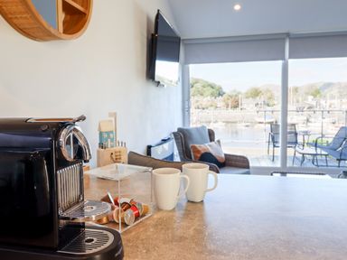 A coffee machine with cups on a countertop and a chair near large windows overlooking a marina at Sea Breeze in Porthmadog