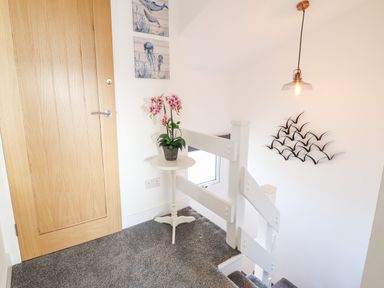 A staircase landing with a wooden door a small round table with a potted pink flower a window wall art of birds and a hanging light at Sea Breeze in Porthmadog