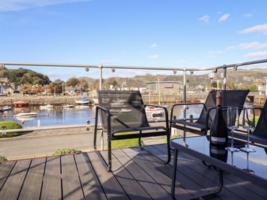 A balcony with black chairs and a glass table with a bottle and two glasses overlooking a harbor with boats at Sea Breeze in Porthmadog