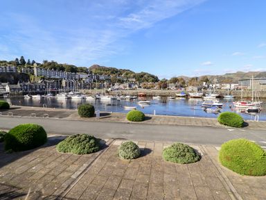 A harbor with boats docked near a waterfront road and bushes with houses and hills in the background at Sea Breeze in Porthmadog