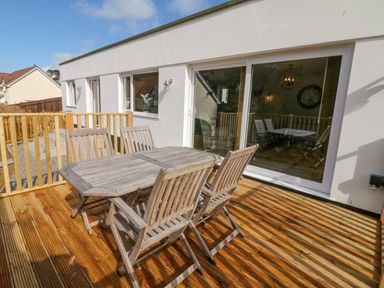 A wooden outdoor table with four chairs on a wooden deck outside a white building with sliding glass doors at Robins Downs in Ilfracombe