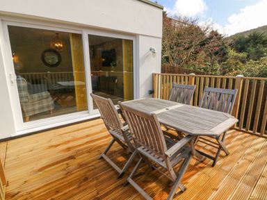 A wooden deck with a wooden table and four chairs outside a house with sliding glass doors at Robins Downs in Ilfracombe