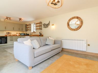A living room with a gray sofa pillows a wooden framed circular mirror kitchen cabinets and shelves in the background at Robins Downs in Ilfracombe