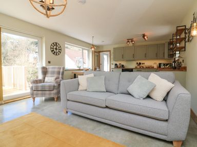 A living room with a gray sofa and plaid armchair next to a sliding glass door with a kitchen in the background at Robins Downs in Ilfracombe