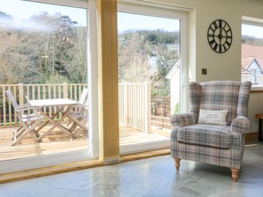 An armchair with a cushion next to large sliding glass doors leading to a wooden deck with a table and chairs at Robins Downs in Ilfracombe
