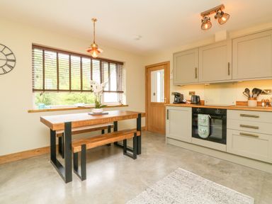 A kitchen with a wooden dining table and bench under a window with blinds and gray cabinets with appliances at Robins Downs in Ilfracombe