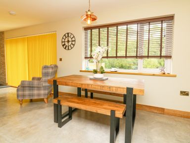 A dining area with a wooden table and bench next to a window with blinds and a plaid armchair near yellow vertical blinds at Robins Downs in Ilfracombe