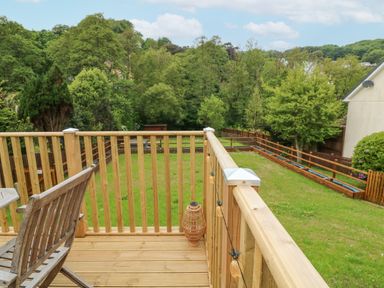 A wooden deck with a chair and table overlooking a fenced lawn with trees in the background at Robins Downs in Ilfracombe