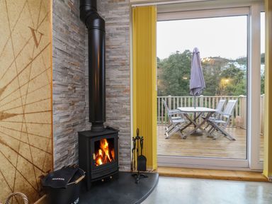A wood burning stove with fire inside next to a large glass door leading to a wooden deck with a table and chairs at Robins Downs in Ilfracombe