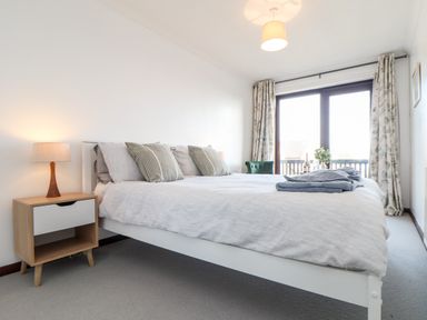 A bedroom with a bed with pillows and a nightstand with a lamp by a window with curtains at Chi an Vre in Newlyn