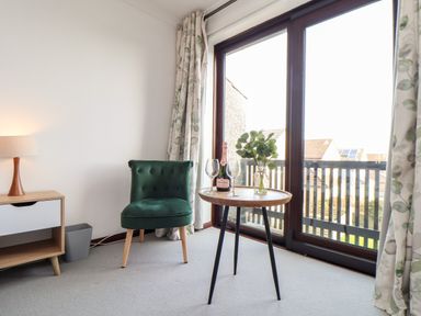 A corner of a room with a green chair a small round table with a bottle glasses and flowers next to a large window with curtains at Chi an Vre in Newlyn