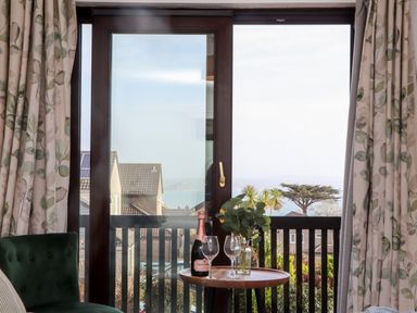 A window with floral curtains overlooking rooftops a tree and the sea with a round wooden table holding a bottle of sparkling wine two wine glasses and a vase with green foliage at Chi an Vre in Newlyn