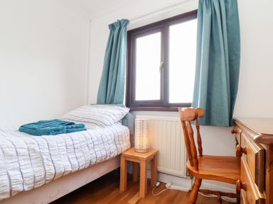 A small bedroom with a single bed next to a window with teal curtains a wooden chair a desk and a bedside table with a lamp at Chi an Vre in Newlyn
