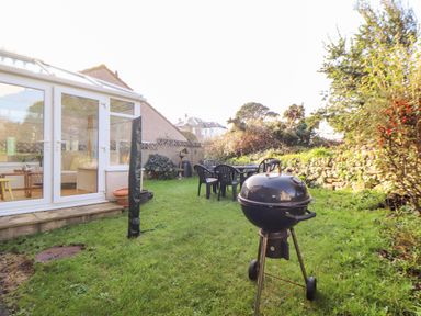 A garden with a charcoal barbecue grill plastic chairs and table next to a conservatory at Chi an Vre in Newlyn