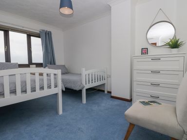 A bedroom with two white beds a chest of drawers with a mirror and a plant and a chair on blue carpet at Chi an Vre in Newlyn