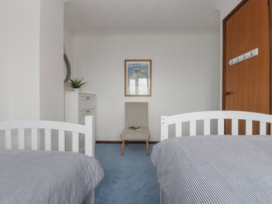 A bedroom with two single beds a gray chair a white chest of drawers a framed picture on the wall and a wooden door at Chi an Vre in Newlyn