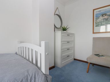 A bedroom with a white bed frame striped bedding a white chest of drawers with a round mirror and a plant on top and a beige chair with a book on it at Chi an Vre Newlyn
