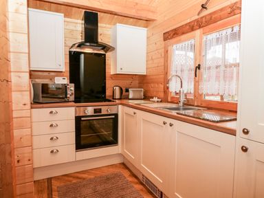 A kitchen with white cabinets a stove microwave kettle sink and window with lace curtains at Ballinorig Lodge in Cleator Moor