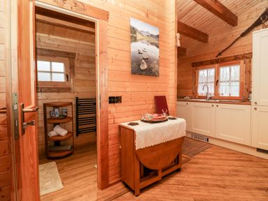 A wooden kitchen with white cabinets a small sink a folding table with a lace cloth and a doorway leading to a bathroom with towels and a wooden shelf at Ballinorig Lodge in Cleator Moor