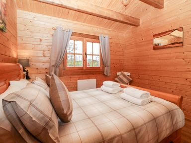 A bedroom with a double bed with plaid bedding and two rolled towels on it next to a window with curtains and a chair at Ballinorig Lodge in Cleator Moor