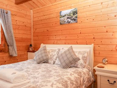 A bedroom with a bed a nightstand with an alarm clock a window with curtains and a landscape picture on the wooden wall at Ballinorig Lodge in Cleator Moor
