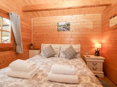 A bedroom with a wooden interior a bed with patterned bedding and pillows bedside tables with a lamp and clock at Ballinorig Lodge in Cleator Moor