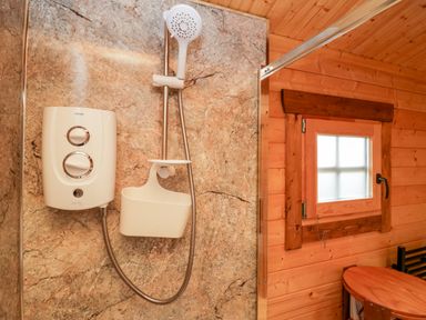A shower area with a water heater unit and showerhead next to a wooden wall with a small window at Ballinorig Lodge in Cleator Moor