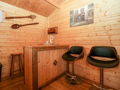 A wooden bar counter with two black bar stools and wooden walls with hanging oversized wooden spoon and fork at Ballinorig Lodge in Cleator Moor