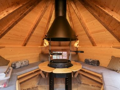 An interior of a wooden cabin with benches and cushions around a central black fireplace at Ballinorig Lodge in Cleator Moor