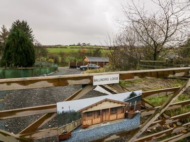 A wooden gate with a sign reading Ballinorig Lodge and a picture of a wooden cabin in a rural area at Ballinorig Lodge in Cleator Moor