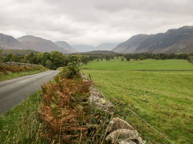 A country road beside a stone wall with green fields trees and mountains in the background at Ballinorig Lodge in Cleator Moor