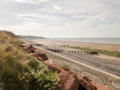 A beach with a sandy shore and rocky cliffside with grassy plants at Ballinorig Lodge in Cleator Moor