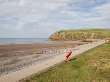 A beach with a concrete walkway grassy area and a hilly cliff in the background at Ballinorig Lodge in Cleator Moor