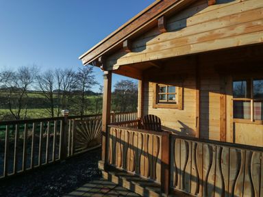 A wooden cabin porch with a chair and wooden railing overlooking trees and green fields at Ballinorig Lodge in Cleator Moor