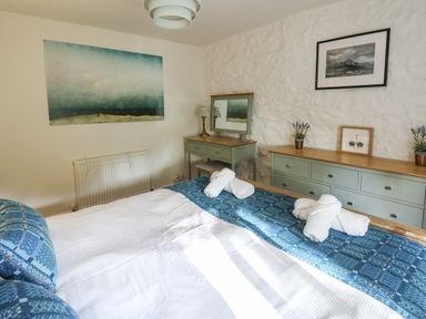 A bedroom with a bed covered in white and blue linens and rolled towels on top a dresser and vanity with a mirror on a stone wall at Ty Len in St Davids