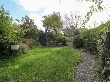 A backyard garden with a wooden picnic table and umbrella on gravel surrounded by grass and bushes at Ty Len in St Davids