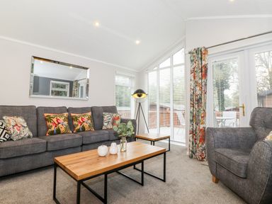 A living room with a gray sofa and armchair a wooden coffee table and side table a floor lamp and large windows with floral curtains at Squirrels Nook Waterside Wood 19 White Cross Bay near Windermere