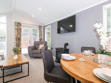 A living room with a round dining table set with plates and glasses a gray armchair a wall-mounted TV and floral curtains at Squirrels Nook Waterside Wood 19 White Cross Bay near Windermere