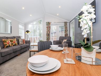 A living room with a gray sofa and armchair with patterned cushions a wooden dining table set with plates bowls and glasses and a white flower plant at Squirrels Nook Waterside Wood 19 White Cross Bay near Windermere