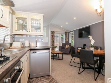 A kitchen area with a sink oven and dishwasher leading to a living room with armchair dining table chairs and a wall-mounted television at Squirrels Nook in Waterside Wood 19 White Cross Bay near Windermere