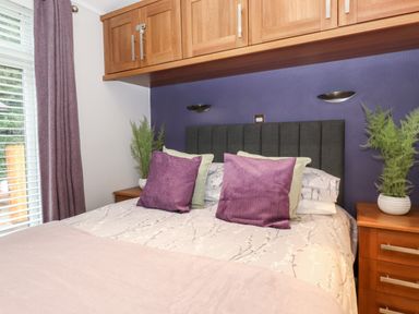 A bedroom with a bed with purple and green pillows bedside tables with plants wooden cabinets above the bed and a window with purple curtains at Squirrels Nook in Waterside Wood 19 White Cross Bay near Windermere